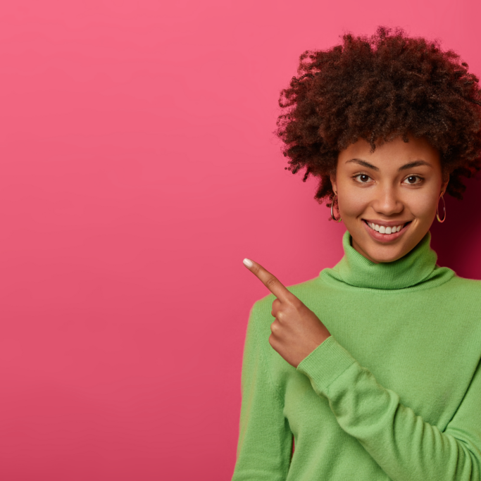 woman with curly hairstyle, healthy skin and white teeth, points on blank place, wears casual green sweater, gives direction